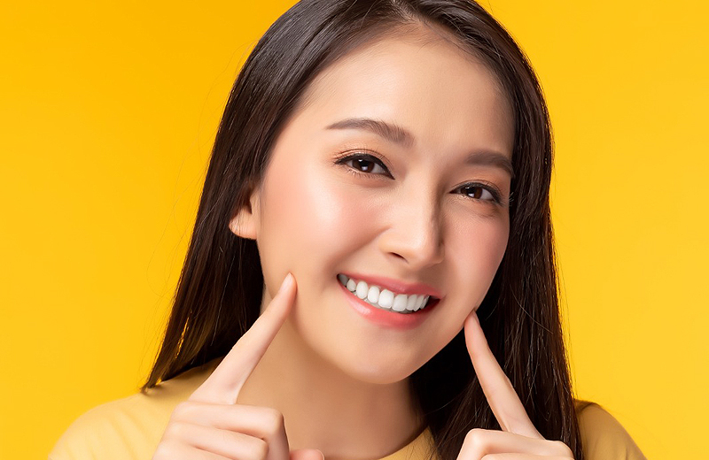Young woman smiling with fingers pointing to cheeks against yellow background for dental practice.