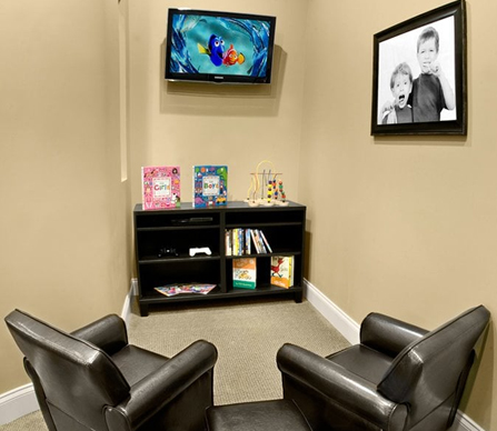 Modern dental office waiting room with two leather chairs, wall-mounted TV, and shelving unit.