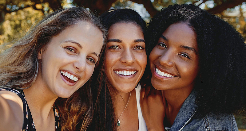 Three smiling women posing together outdoors in natural sunlight, representing dental health and wellness.