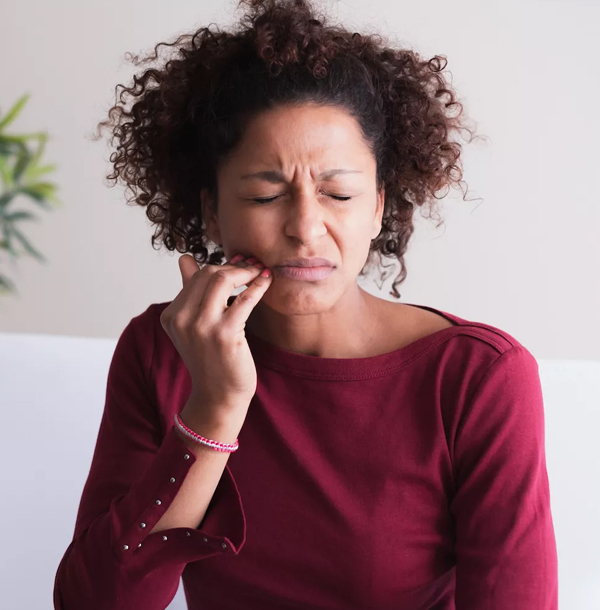 Woman with curly hair in burgundy shirt touching face, appearing concerned or in dental discomfort, indoors.