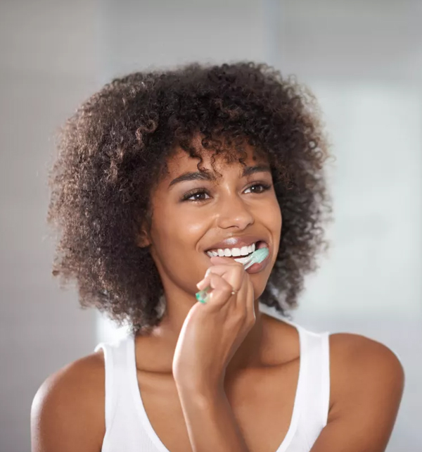 Woman brushing teeth, smiling at camera, white tank top, neutral background, dental hygiene