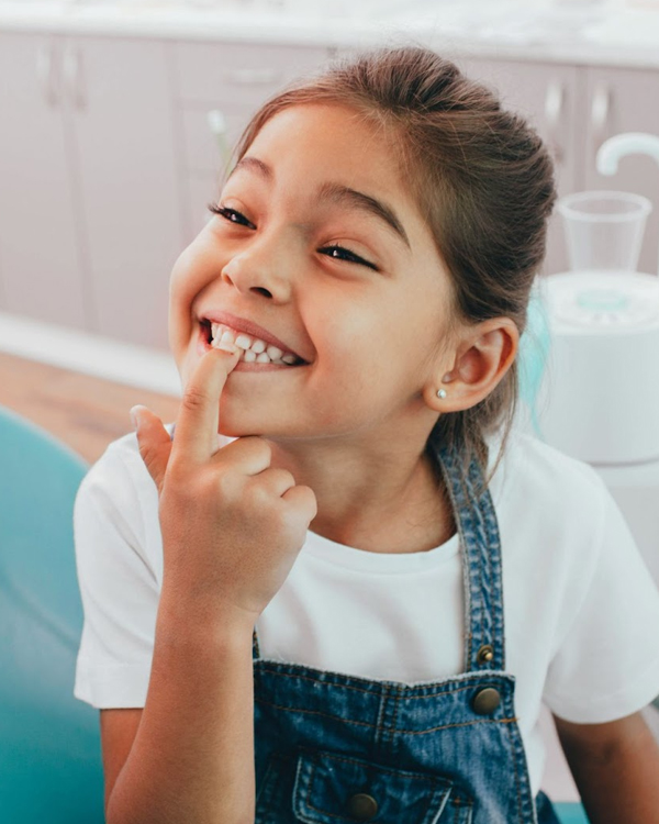 Young girl smiling brightly while pointing to her teeth in a dental office setting.